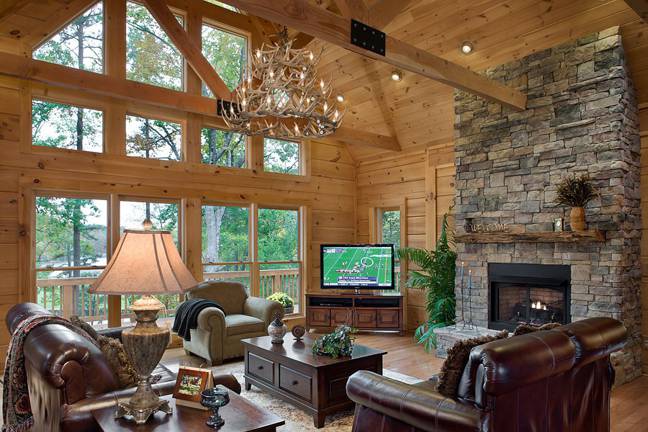 photo of interior shot of log home with soaring ceilings and two story window treatment with door leading to open porch. Fireplace is floor to ceiling stone